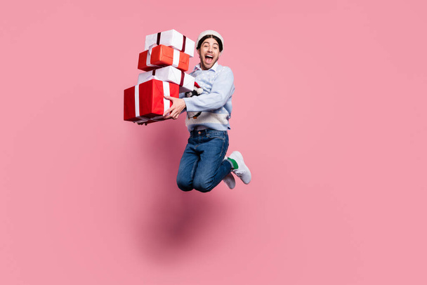 Photo of cute shocked young man dressed blue christmas pullover jumping holding presents pile smiling isolated pink color background - Foto, imagen