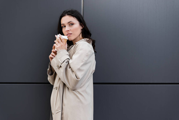 mujer joven en abrigo beige de pie con bebida para llevar cerca de la pared gris - Foto, Imagen