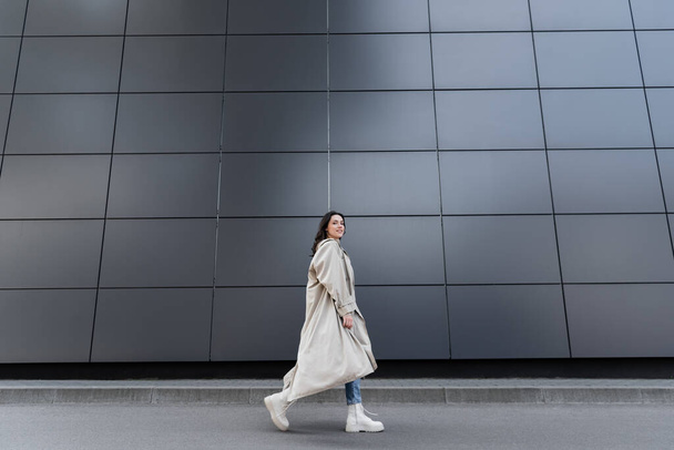 trendy woman in long raincoat and white boots walking along grey wall - Foto, Imagen