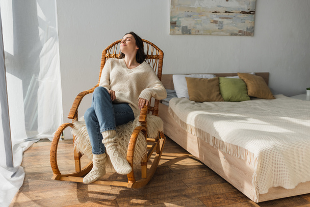 joyful woman in soft sweater and warm socks resting in rocking chair in bedroom - Photo, Image
