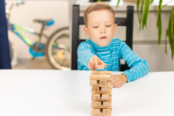 A cute seven-year-old child boy playing Jenga at home at a white wooden table against the background of a light window. Selective focus. Close-up. Portrait - Fotografie, Obrázek