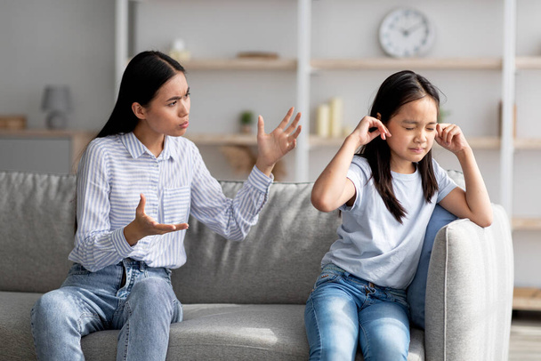 Mutter-Tochter-Konflikt. Junge asiatische Mutter versucht, mit ihrem beleidigten kleinen Kind zu sprechen, sitzt zusammen auf dem Sofa zu Hause - Foto, Bild