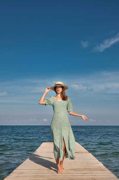 Atraktivní mladá běloška v zelených šatech a slamáku lehce kráčí po dřevěném molu za slunečného dne na pozadí moře. Odpočiňte si a procházejte se po oceánu - Fotografie, Obrázek