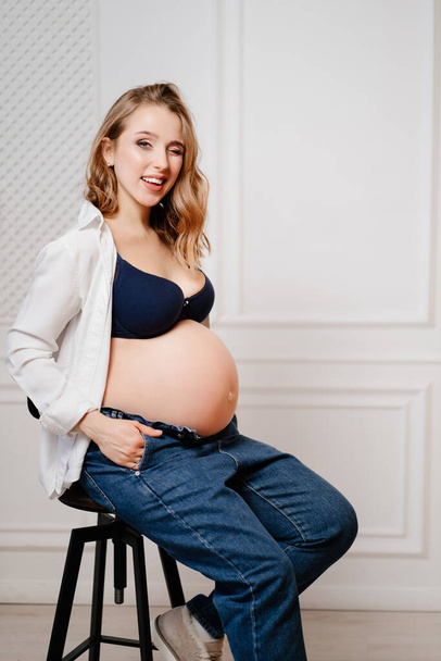 привлекательная беременная женщина в белой рубашке с голым животом сидит на стуле. - Фото, изображение