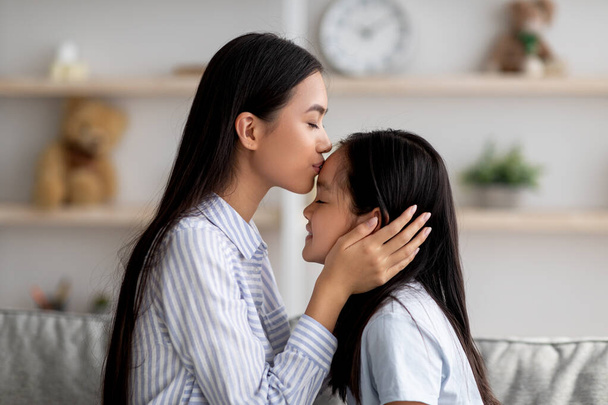 Матери любят. Любящая азиатская мама целует свою дочь в лоб, проводя время вместе дома, вид сбоку - Фото, изображение