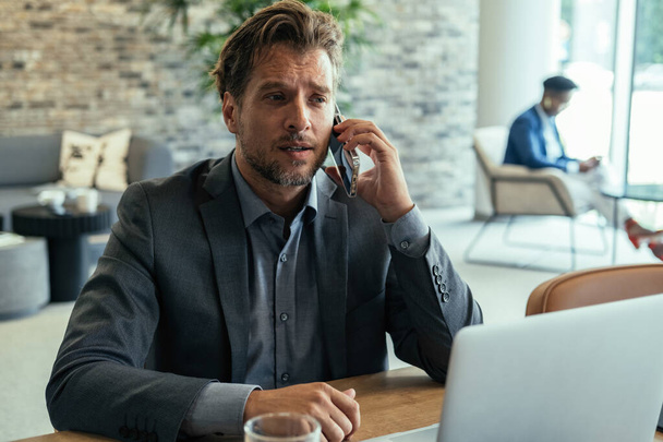 Poważny biznesmen rozmawiający przez telefon komórkowy w pracy. Zaniepokojony niezadowolony biznesmen kłócący się przez telefon siedząc przy biurku i pracując na laptopie w biurze. - Zdjęcie, obraz