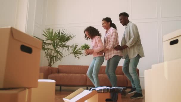 Glückliche afrikanisch-amerikanische Dreierfamilie tanzt mit heimischer Pflanze zwischen vielen Pappkartons in neuer eigener Wohnung - Filmmaterial, Video