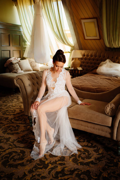 morning of bride. beautiful woman in lace robe sitting on couch in room.  - Фото, изображение