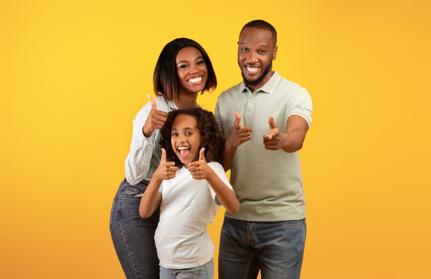 ¡Eh, tú! Emocionados padres negros y su hija señalando con los dedos a la cámara, de pie sobre el fondo amarillo - Foto, imagen