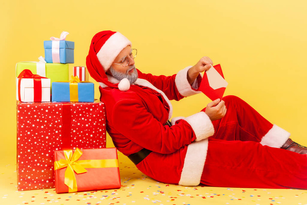 Side view of elderly man with gray beard in santa claus costume sitting on floor near present boxes, reading letter with Christmas congratulations. Indoor studio shot isolated on yellow background. - Foto, Imagen