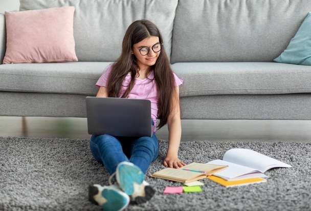 Focused Indian teen girl sitting on floor with laptop and study materials, learning online from home, free space - Foto, afbeelding