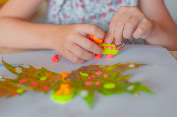 Evde çocuk yaratıcılığı. Küçük kız, yaprakların arka planında parlak plastikten bir oyuncak yapıyor. - Fotoğraf, Görsel