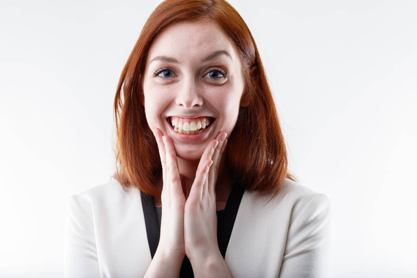 Jonge vrouw kijkt in verrukking en opwinding naar de camera met haar handen op haar wangen en een levendige tand glimlach in een bijgesneden portret op wit - Foto, afbeelding