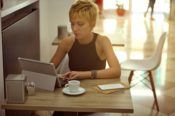 Retrato de una hermosa joven con corte de pelo corto y moderno trabajando en su tablet PC con teclado extraíble junto a una taza de café en su escritorio - Foto, Imagen