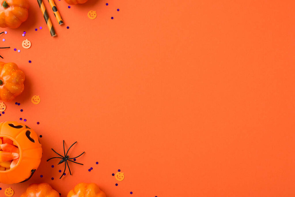 Bovenaanzicht foto van Halloween decoraties pompoen mand met snoep maïs rietjes spin en violette pailletten op geïsoleerde oranje achtergrond met lege ruimte - Foto, afbeelding