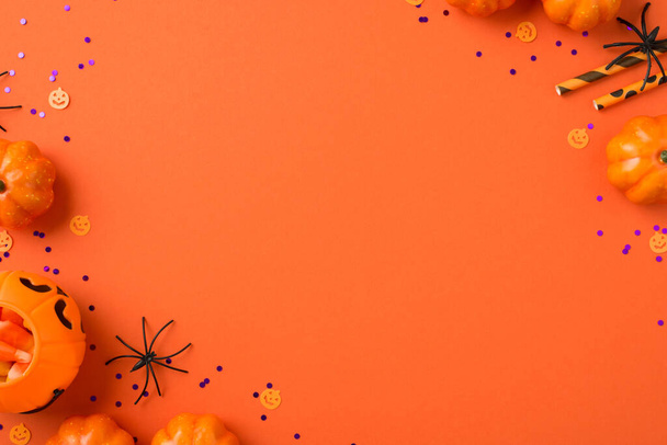 Top view photo of halloween decorations pumpkin basket with candy corn straws spiders and violet sequins on isolated orange background with copyspace - Foto, Imagen