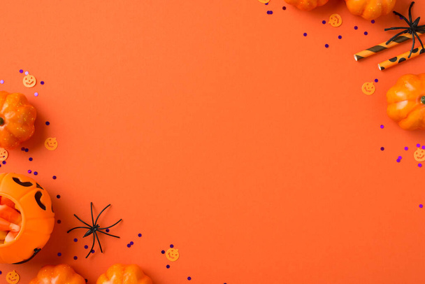 Foto von oben von Halloween-Dekorationen Kürbis Korb Bonbons Mais Strohhalme Spinnen und violettes Konfetti auf isoliertem orangefarbenem Hintergrund mit Kopierraum - Foto, Bild