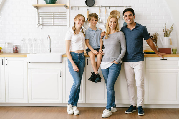 Portrait of happy family with teenage daughter and little son standing on wooden floor in modern kitchen and looking at camera while new renovated home together. Full-length image of family at home. - Photo, Image