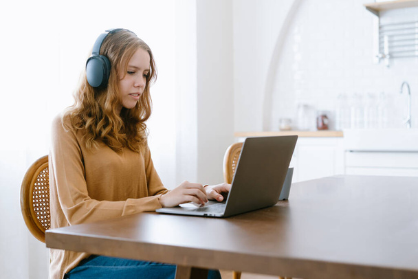 Szczęśliwa nastolatka w słuchawkach patrzy na laptop e-learning ucząc się samotnie w domu. Nastoletnia studentka słuchająca kursu audio lub muzyki podczas odrabiania lekcji. Koncepcja kształcenia zdalnego. - Zdjęcie, obraz