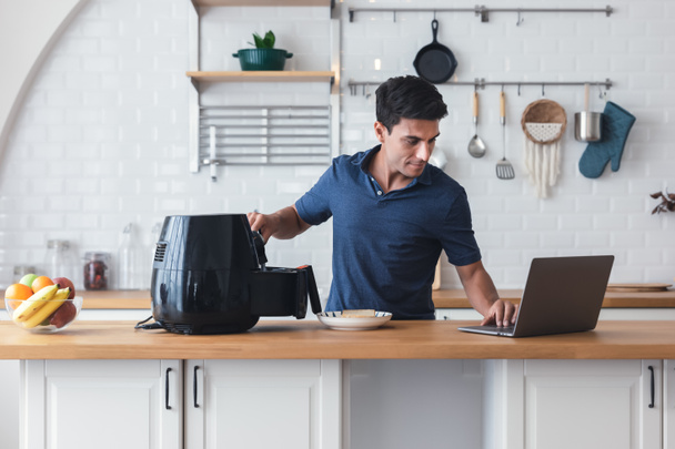 Happy young man standing at kitchen table and cooking toast bread by Air Fryer machine, watching culinary TV show online on laptop. New normal work lifestyle at home. Technology smart cooking device. - Foto, Imagen