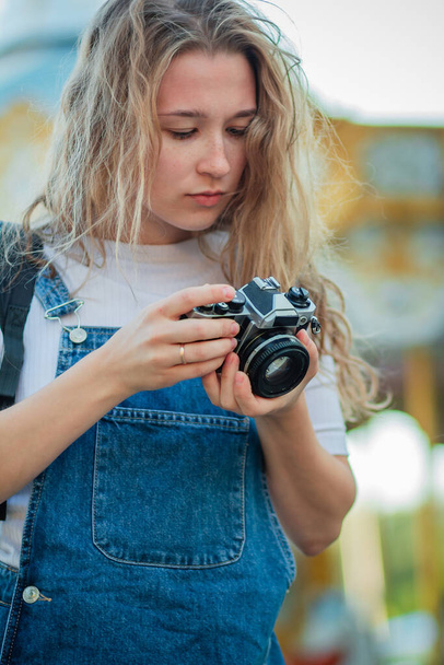 Mladá turistka v džínových šatech stojí s kamerami v zábavním parku. Teen dívka v moderním oblečení fotí na pozadí kolotoče - Fotografie, Obrázek