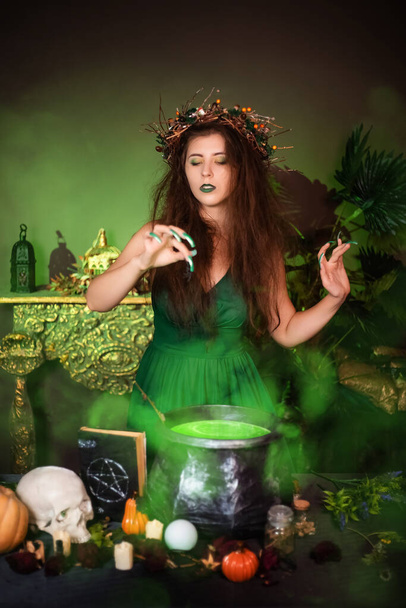 Una bruja con el pelo desaliñado y las uñas largas con una corona de ramas secas en la cabeza, vestida con un vestido verde, prepara una poción mágica de pie junto a un caldero y una mesa con ingredientes. - Foto, imagen