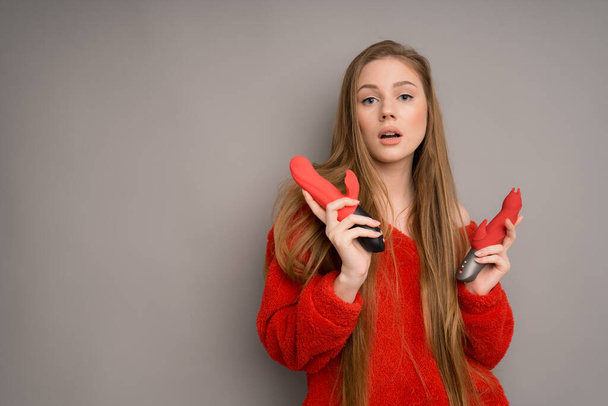 A pretty girl in a sweater stands with her mouth open and holds two red dildos or vibrators on a gray background. - Photo, Image