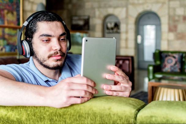 A side view of a young attractive man sitting on a comfortable couch and reading an e-book on a tablet - Foto, immagini