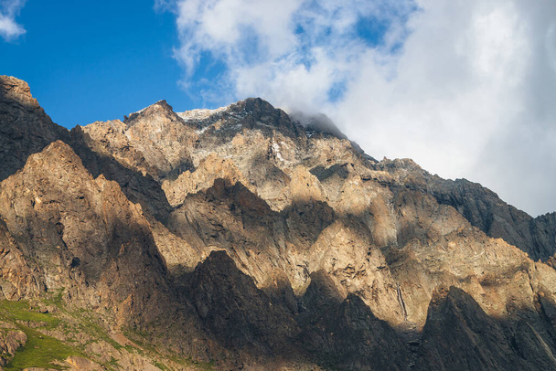Γραφικό ορεινό τοπίο με μεγάλους βράχους στο χρυσό φως του ήλιου και χαμηλά σύννεφα. Φοβερός βραχώδης τοίχος με αιχμηρή κορυφή σε χρυσό ήλιο. Πολύχρωμο ηλιόλουστο τοπίο με ψηλό βραχώδες βουνό με μυτερή κορυφή. - Φωτογραφία, εικόνα