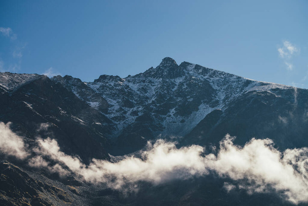 Wspaniały widok na skalistą ścianę górską ze śniegiem nad chmurami i szczytowym szczytem. Malowniczy krajobraz górski ze szpiczastym szczytem i niskimi chmurami na skałach. Minimalna sceneria alpejska z ostrym, skalistym wierzchołkiem. - Zdjęcie, obraz