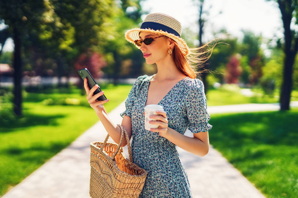 Young female in dress straw hat, fashionable sunglasses surfing or chatting by phone and drinking coffee while stroll outside in summer green park Holding straw handbag and coffee - Foto, Imagem