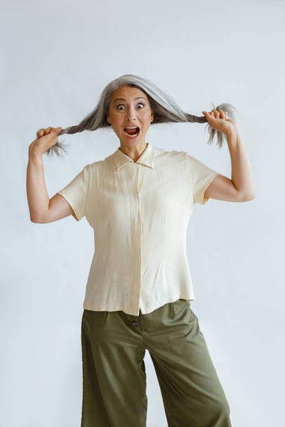Shocked Asian woman holds hoary hair locks on light grey background - Foto, afbeelding