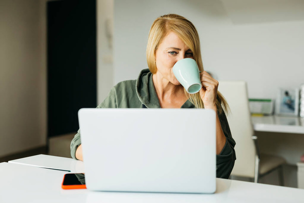 Donna bionda di mezza età che usa un computer portatile in soggiorno e beve caffè su una tazza - Foto, immagini