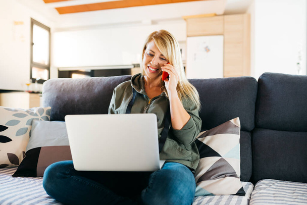 Lächelnde blonde Frau mittleren Alters mit Laptop und einem Gespräch auf einem roten Smartphone auf dem Sofa eines Wohnzimmers - Foto, Bild