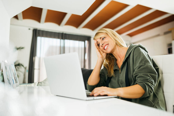 Középkorú, mosolygós szőke nő laptoppal és videóhívással a nappaliban - Fotó, kép