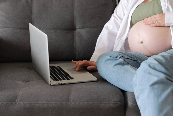 Evden internet üzerinden çalışan çekici hamile bir genç bayan. Kadın ellerini dizüstü bilgisayarın üzerine koy ve karnında büyük bir hamilelik var.. - Fotoğraf, Görsel