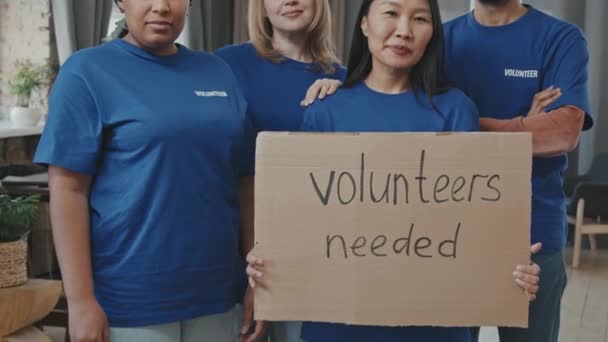 Odchyl średni portret czterech uśmiechniętych, wieloetnicznych wolontariuszek i wolontariuszek patrzących w kamerę z kartonowymi wolontariuszkami Potrzebny znak - Materiał filmowy, wideo