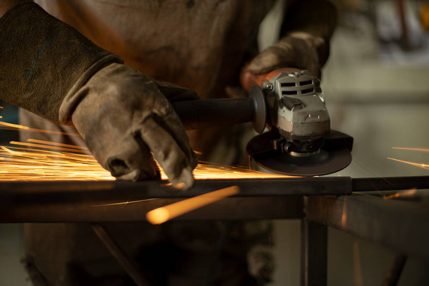 Metal processing with a grinder. Sparks from the tool. A man in the workshop wearing gloves with a grinder in his hand. Sparks from grinding steel. - 写真・画像
