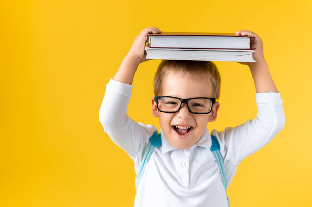 Funny Preschool Child Boy in Glasses with Book on Head and Bag on Yellow Background Copy Space. Happy Smiling Kid Go Back to School, Kindergarten. Success, Motivation, Winner Genius, Superhero concept - Foto, Imagem