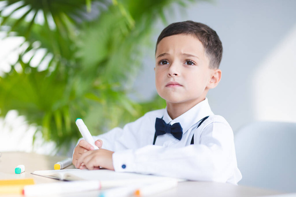 Elementary school kid is sitting at table, serious, thinking. White room with houseplant, desk with school supplies. Intelligent child is focused. Concept of school, learning, education, achievement - Фото, зображення