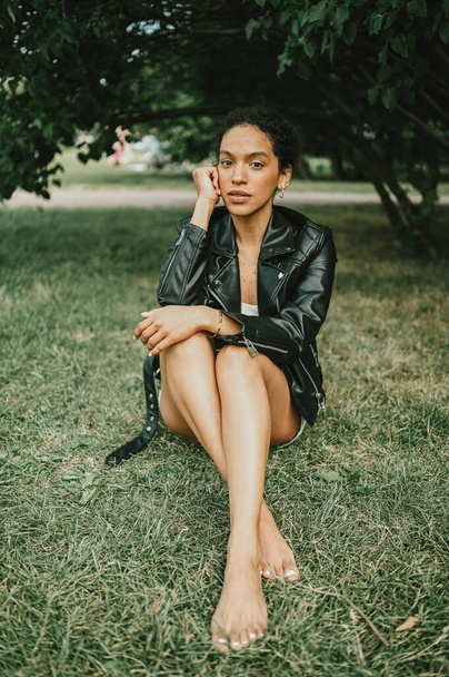 Fashion portrait of attractive confident young natural beauty African American woman with afro hair in black leather jacket posing in nature park in green foliage - Φωτογραφία, εικόνα