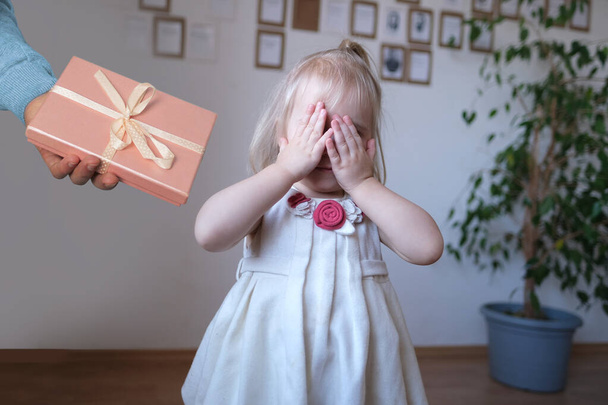 small child in a white dress, baby, blonde girl mischievously covered her face with her palms, hand holds out a gift, parenting concept, family everyday life - Фото, изображение