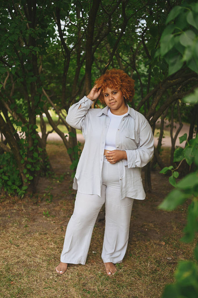 Fashion portrait of young naturally beautiful plus size African American woman in light trouser suit with afro red hair posing in nature parkland in green foliage. Diversity and body positive concept. - 写真・画像