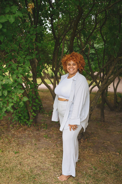 Fashion portrait of young naturally beautiful plus size African American woman in light trouser suit with afro red hair posing in nature parkland in green foliage. Diversity and body positive concept. - Fotó, kép