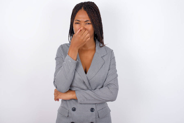 Joven mujer de negocios afroamericana con trenzas sobre pared blanca, sosteniendo su nariz debido a un mal olor. - Foto, Imagen