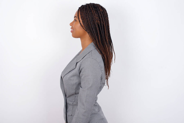 Profil der lächelnden jungen afrikanisch-amerikanischen Geschäftsfrau mit Zöpfen über der weißen Wand mit gesunder Haut, hat besinnlichen Ausdruck, bereit für einen Spaziergang im Freien. - Foto, Bild