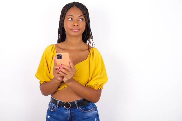 Portrait of  african american woman wearing yelllow top and jeans  with dreamy look, thinking while holding smartphone. Tries to write up a message. - Valokuva, kuva