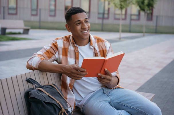 Lachende Afro-Amerikaanse student studeren, leren van taal zitten op de campus, onderwijs concept. Jonge knappe man boek lezen, ontspannen in het park - Foto, afbeelding