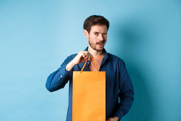 Ευτυχισμένος νεαρός άνδρας δείχνει πορτοκαλί τσάντα ψώνια και κλείσιμο του ματιού, συνιστώντας εκπτώσεις κατάστημα, στέκεται σε μπλε φόντο - Φωτογραφία, εικόνα