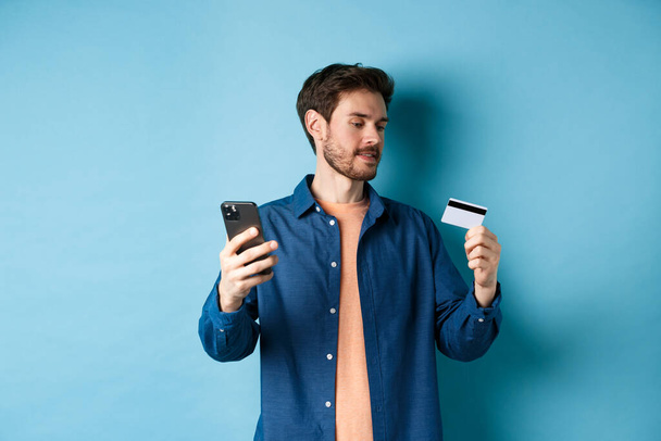 Sähköisen kaupankäynnin käsite. Mies maksaa verkkomaksun, lukee luottokortin numeron ja pitää matkapuhelinta, seisoo sinisellä taustalla - Valokuva, kuva